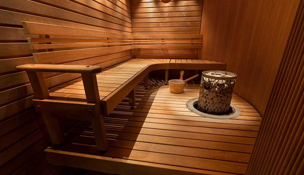 The wooden interior of a sauna, a large L-shaped bench around a stone firepit
