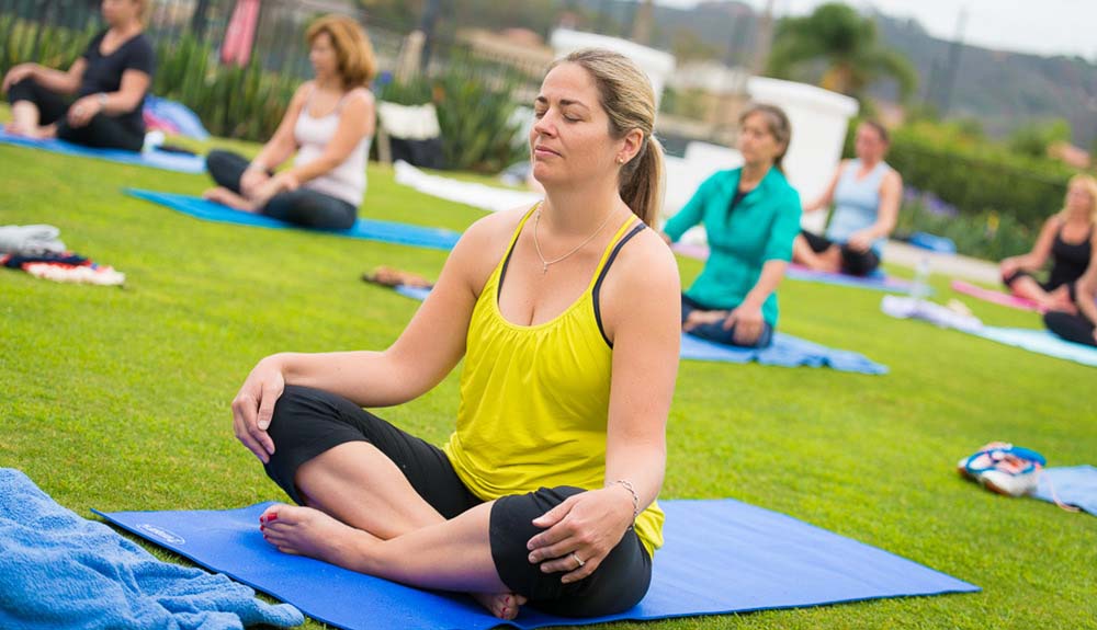 A woman closes her eyes as she sits cross-legged on a yoga mat in the grass, other class participants can be seen in the same position on their own yoga mats behind her
