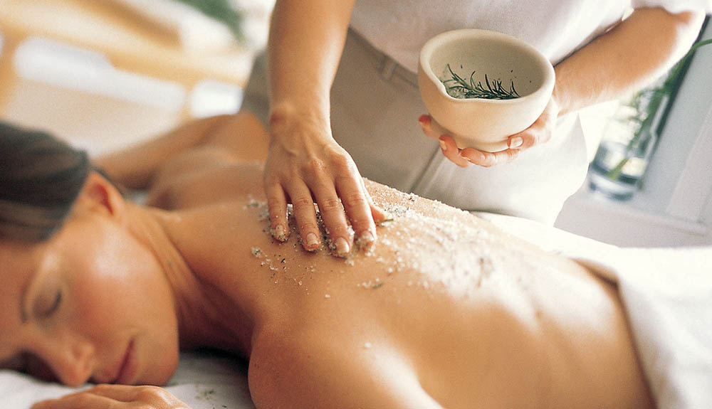A woman enjoys a spa experience with her eyes close and a relaxed look on her face as a scrub is applied to her back