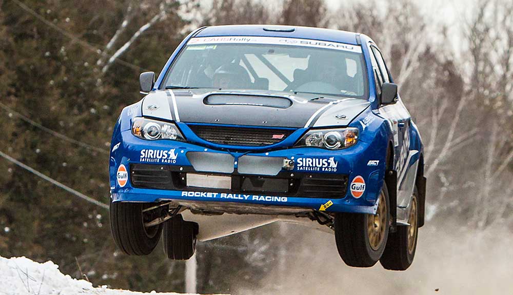 Blue Mitsubishi EVO VI flyng off a snowy race track, wheels in the air as it speeds along the track