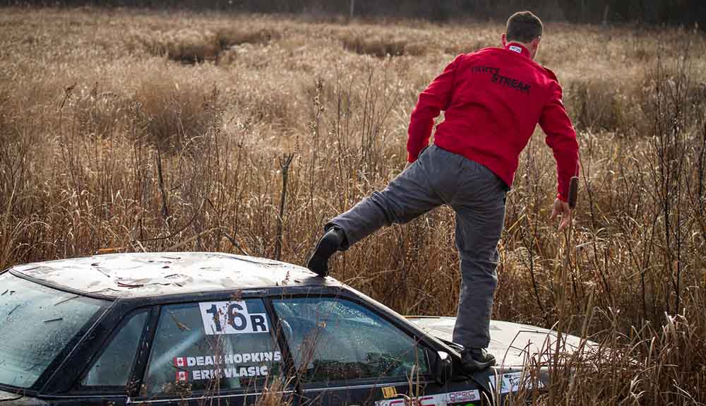 Man in grey pants and red sweatshirt stands on the hood of a car parked amongst tall grasses