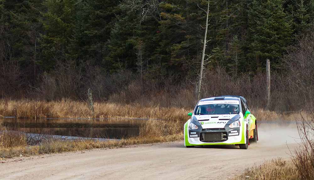 White race car with fluorescent green and yellow stripes speeds down a dusty road beside a small pond
