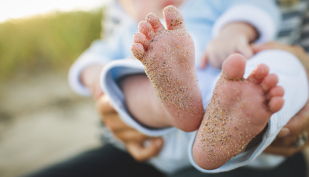 Sandy baby feet seen in the foreground, hands holding baby wearing a light blue playsuit