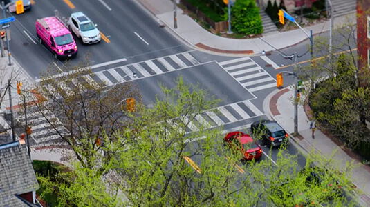 Aerial view of a vibrant urban intersection during daytime, featuring clearly marked pedestrian crosswalks, traffic signals, and several vehicles including a distinctive pink van with white graphics. The streets are lined with green trees and bordered by residential and commercial buildings. Sidewalks are well-maintained, and the scene reflects a blend of city infrastructure and natural elements, suggesting spring or early summer.