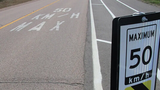 A multi-lane road with a posted speed limit sign reading 'MAXIMUM 50 km/h' and matching road markings that say '50 KM/H MAX' in large white letters. The road includes a bike lane or shoulder lane marked by white lines, emphasizing traffic regulations and safety measures.