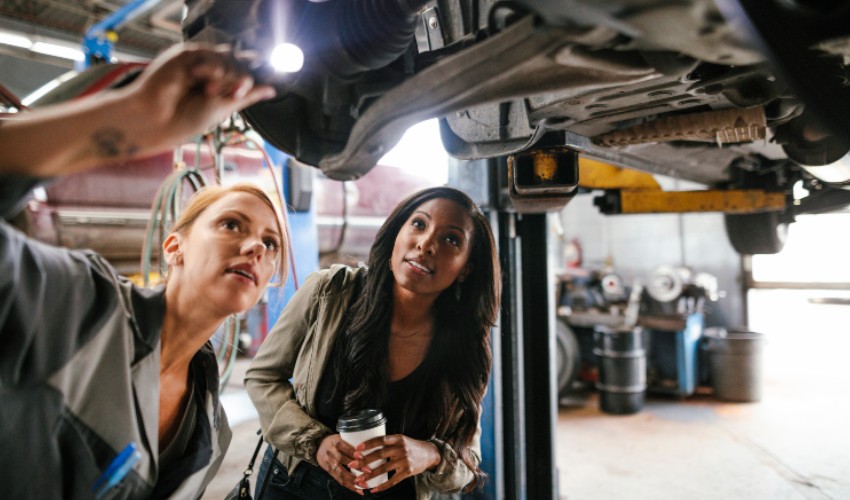 Female mechanic showing a female customer the underbody of a car on a lift in a garage.  