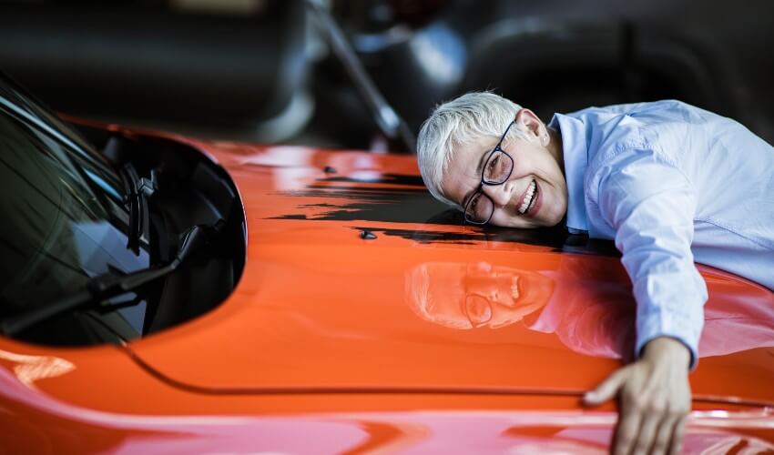 Hip older woman leaning over to hug front of orange car