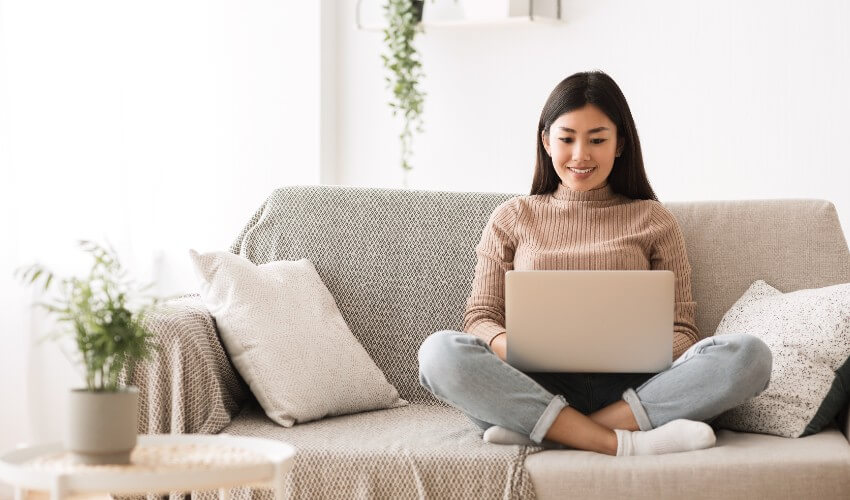 Young Asian woman sitting cross legged on couch in livingroom working on laptop.