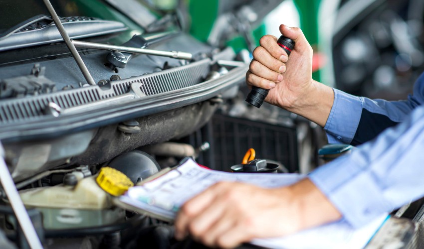 Man with a flashlight and checklist inspecting under the hood of a used car.