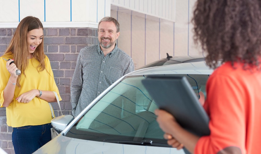 A young woman, sales agent and parent stand by as she admires her new used car purchase.