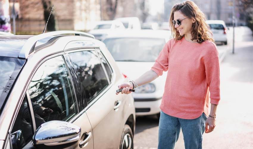 Happy cheerful young woman driver in sunglasses opens her car with a remote key on the street in the city
