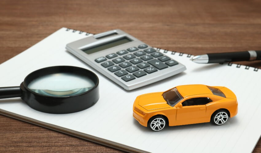 Yellow toy car, magnifying glass, calculator, pen and notebook sitting on wooden desk.