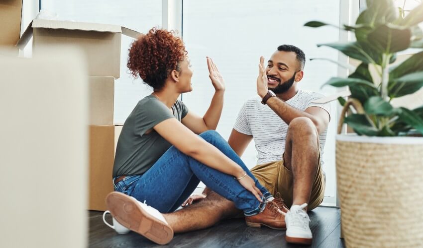Young African-American couple high-fiving each other while sitting on floor of new home, surrounded by packing boxes.