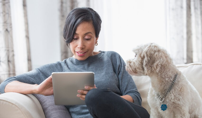 African American woman seated on couch looking at digital tablet, while her white furred dog watches her.