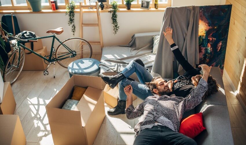 Young couple relaxing on couch in messy apartment living room, surrounded by unpacked moving boxes, bike and wrapped artwork.