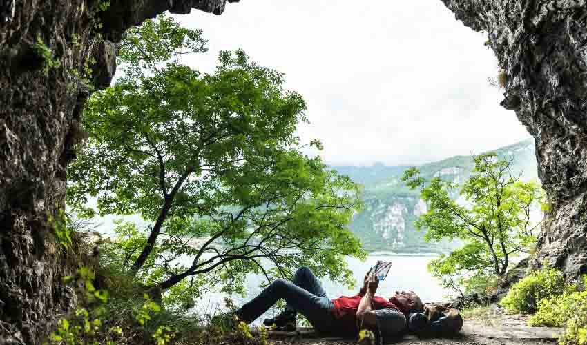 Male hiker outside a cave laying down reading a book.
