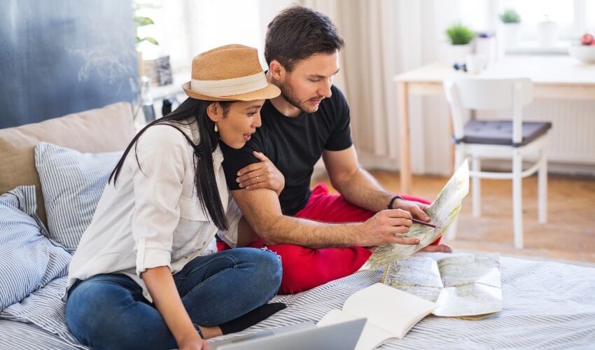Young mixed ethnicity couple sitting on bed with laptop, looking at map and planning trip.