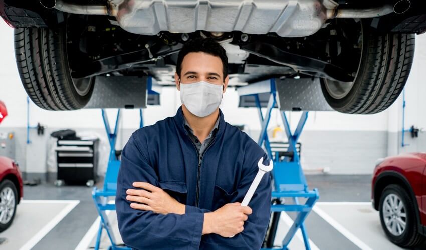 Mechanic wearing a mask standing in front of a car on a hoist. 