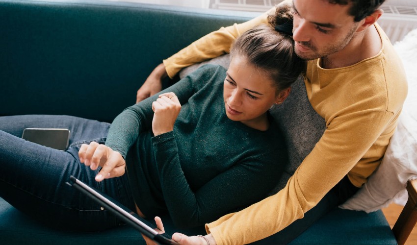 Young couple relaxing on a couch, surfing for travel destinations on a tablet.