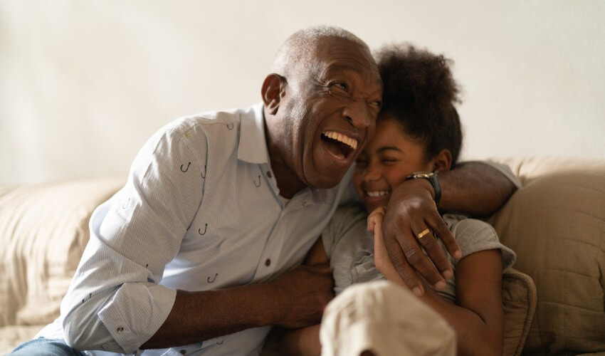 Grandfather laughing while hugging granddaughter on couch.