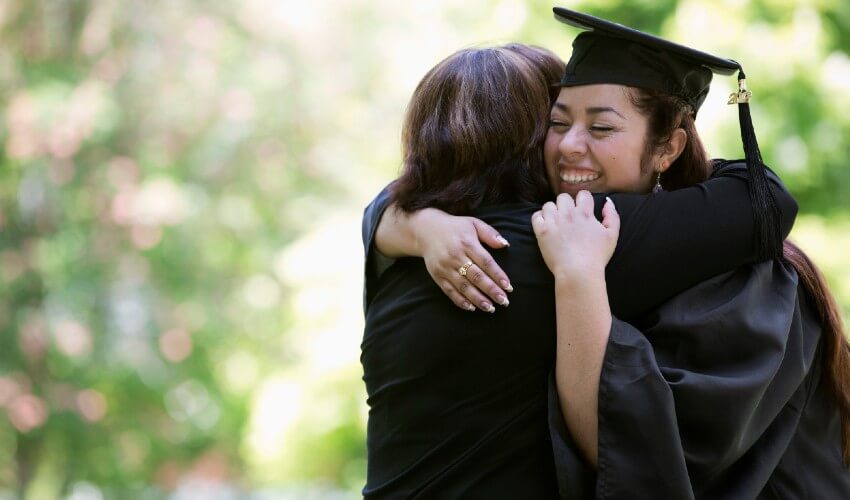 University graduate in cap and gown, hugging her mom outdoors beneath a tree.
