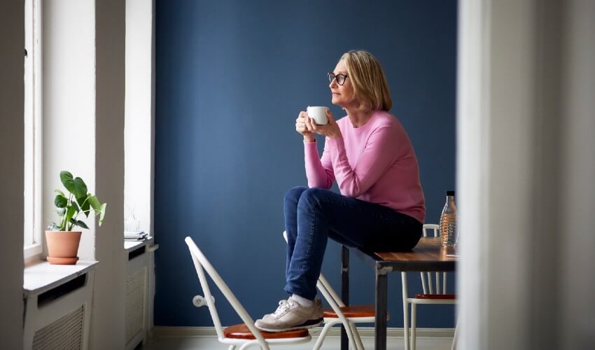 Middle age woman holding coffee cup and sitting on table, staring out window pensively.