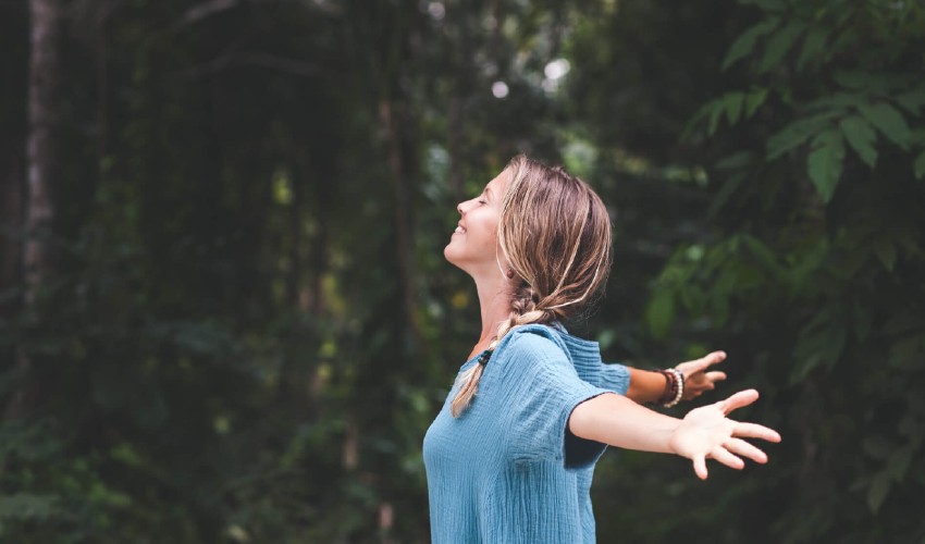 Smiling and peaceful woman standing with outstretched arms in the forest. 