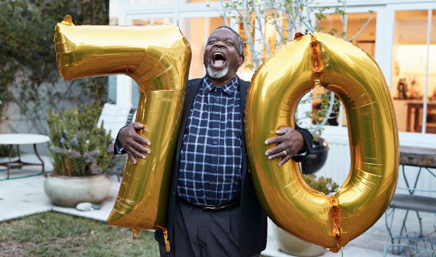 Man in his backyard, laughing as he holds balloons reading age seventy. 