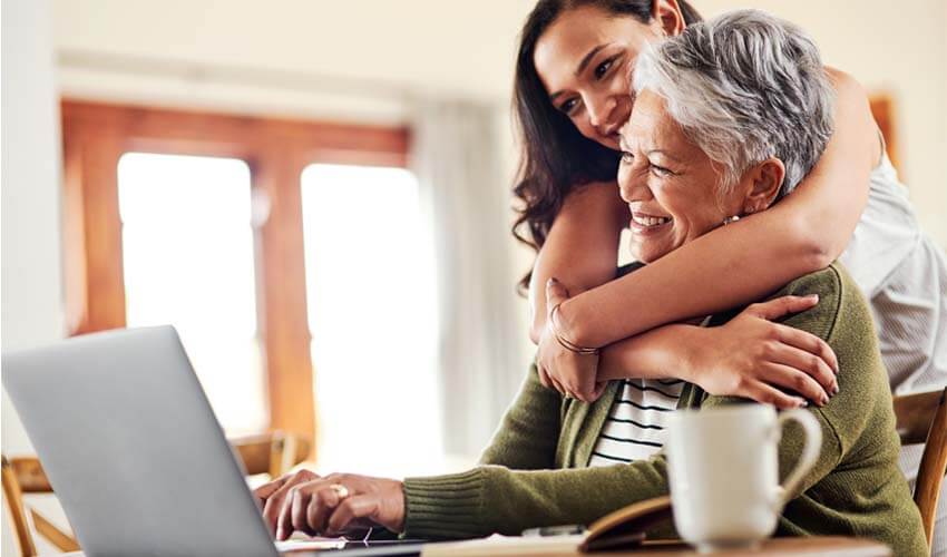 Young woman hugging her Grandmother who is seated in front of a laptop.