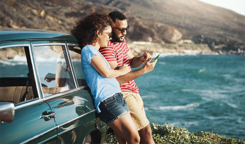 Couple leaning on car by shore looking at phone