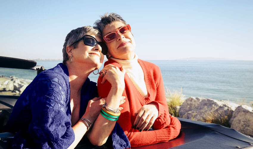 Two mature ladies leaning on car by shore