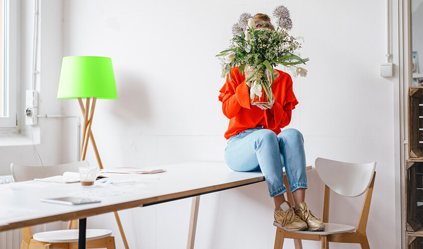 Woman sitting on table hiding behind vase of flowers.