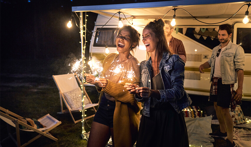 Four adults laughing outside a camper at night with two of them holding glowsticks.
