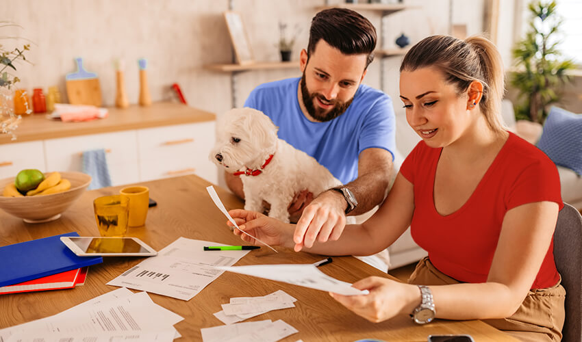 A man and a woman sitting at a table with their small white dog looking at paperwork.