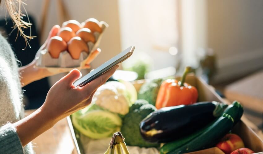 Woman using smartphone while checking fresh fruits and vegetables delivery box at home.