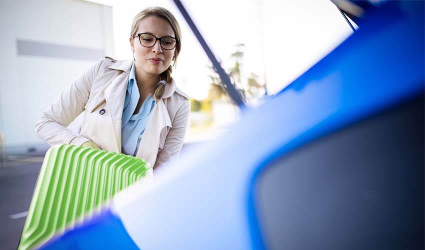 Woman putting green suitcase in trunk of blue car