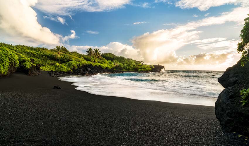 Waves lapping on a deserted black sand beach in Maui, beneath a cloud-strewn sky.