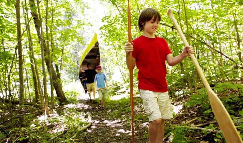 Teenaged boy carrying canoe paddles in forest. Dad and younger boy follow behind carrying yellow canoe and other paddles.