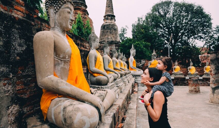 Woman holding young daughter on shoulders as they both look at row of Buddhas in Thailand.
