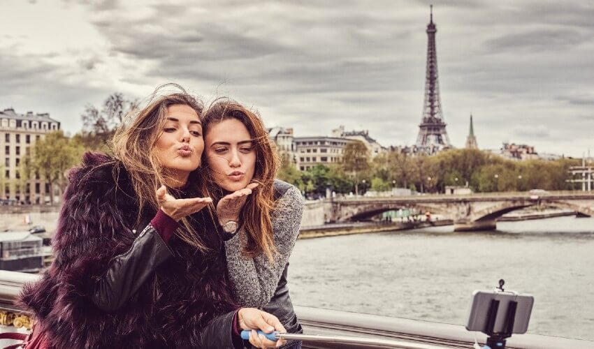 Two young girls blowing kisses while posing for a selfie with the Eiffel Tower in background.