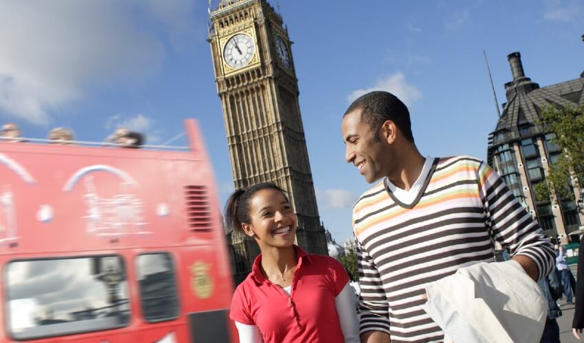 Young couple walking in London in front of Big Ben, with red double decker bus in background.