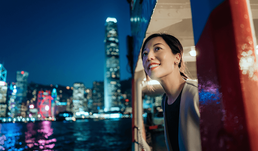 Young woman looking out through a ferry window, enjoying a night illuminated city.