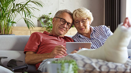Aged couple with a mans leg in a cast, sitting on a couch looking at a tablet