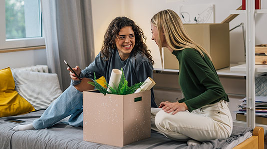 Two woman sitting on a bed with a box, both looking at a phone.