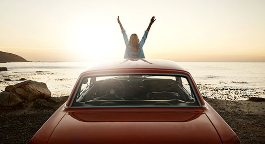 Young female sitting on hood of car with hands raised excited to enjoy the sunset and view of coast.