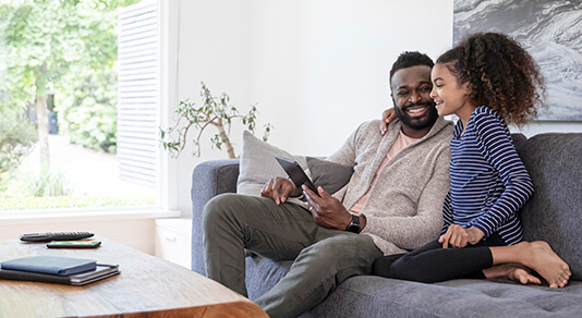 A man smiles with his daughter on the couch as they interact with a digital tablet