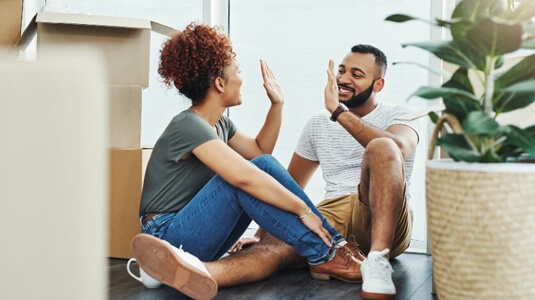 Young couple high fiving each other while sitting on floor surrounded by packing boxes