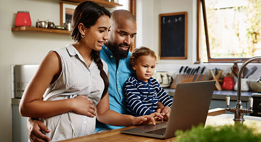 A family of three gathered around a kitchen counter looking at a laptop screen.