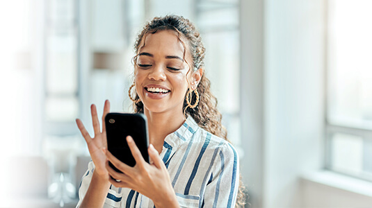 A smiling woman with curly hair looks at her smartphone and gestures with her hand, standing indoors near a bright window.