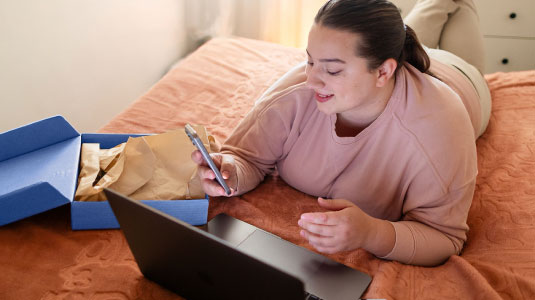A woman is on a bed, looking at her phone, with a laptop and an open box beside her